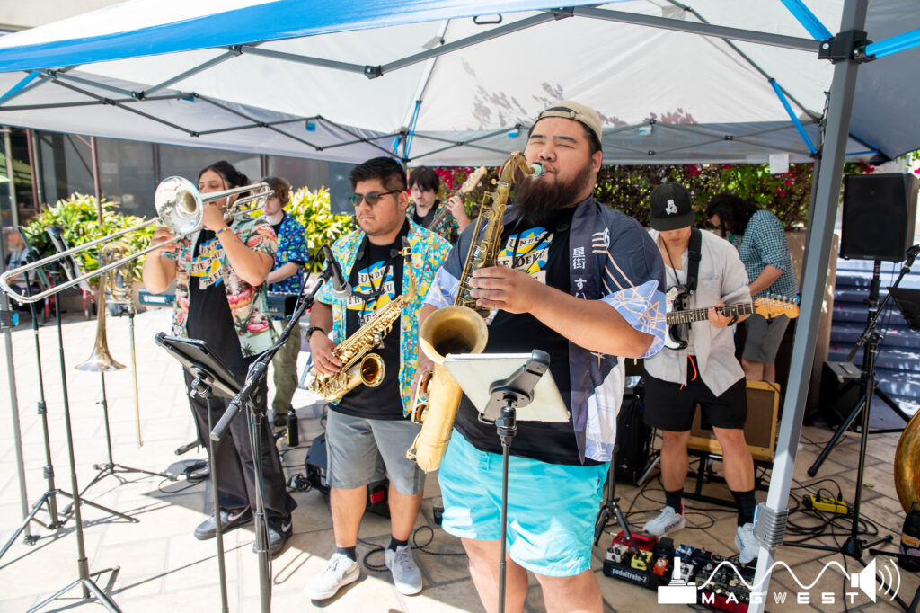 A band comprised of guitars, a trombone, and saxophones are decked out in Hawaiian shirt finery and playing tunes under a shaded canopy by the pool.