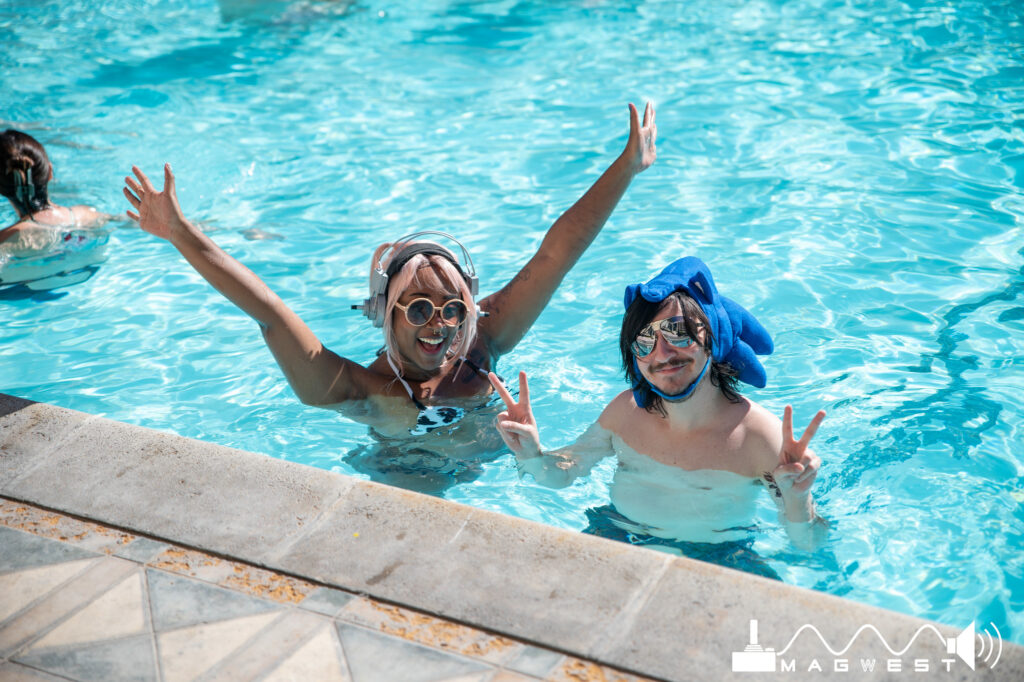 Two people are in the pool wearing sunglasses. One is wearing a headset and has their arms raised up and the other is wearing a sonic hat and giving peace signs.