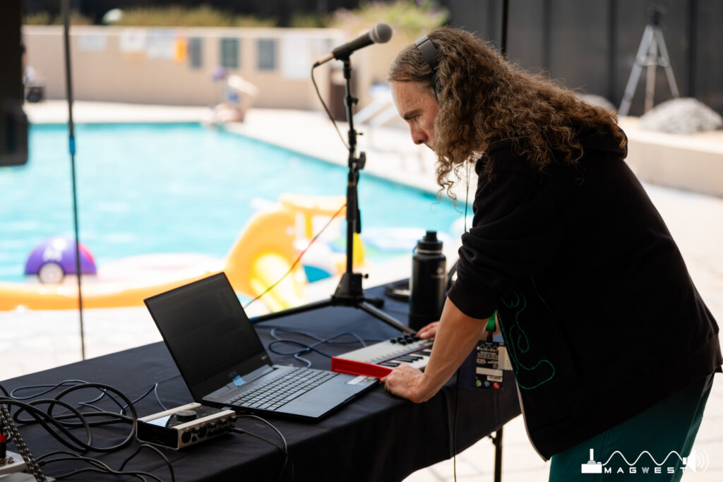 A person with long curly brown hair wearing a black hoodie looks at a laptop. A small piano and microphone is in front of them. The pool with several floaties is in the background.